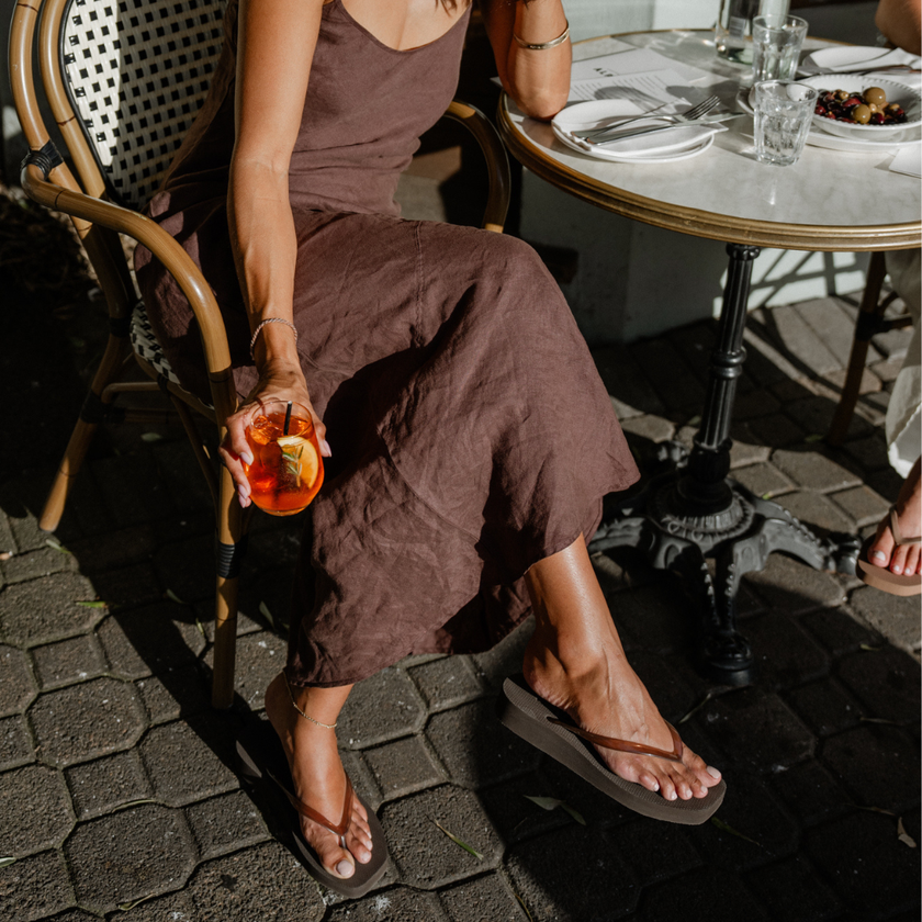 Woman in a brown dress seated at a café wearing Espresso Jelly Tapered Platform flip flops by Sandos