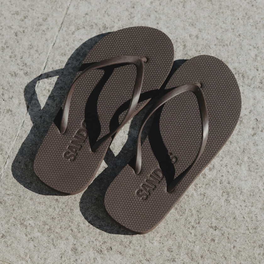 Pair of dark brown Truffle Boyfriend Wide flip flops by Sandos on concrete